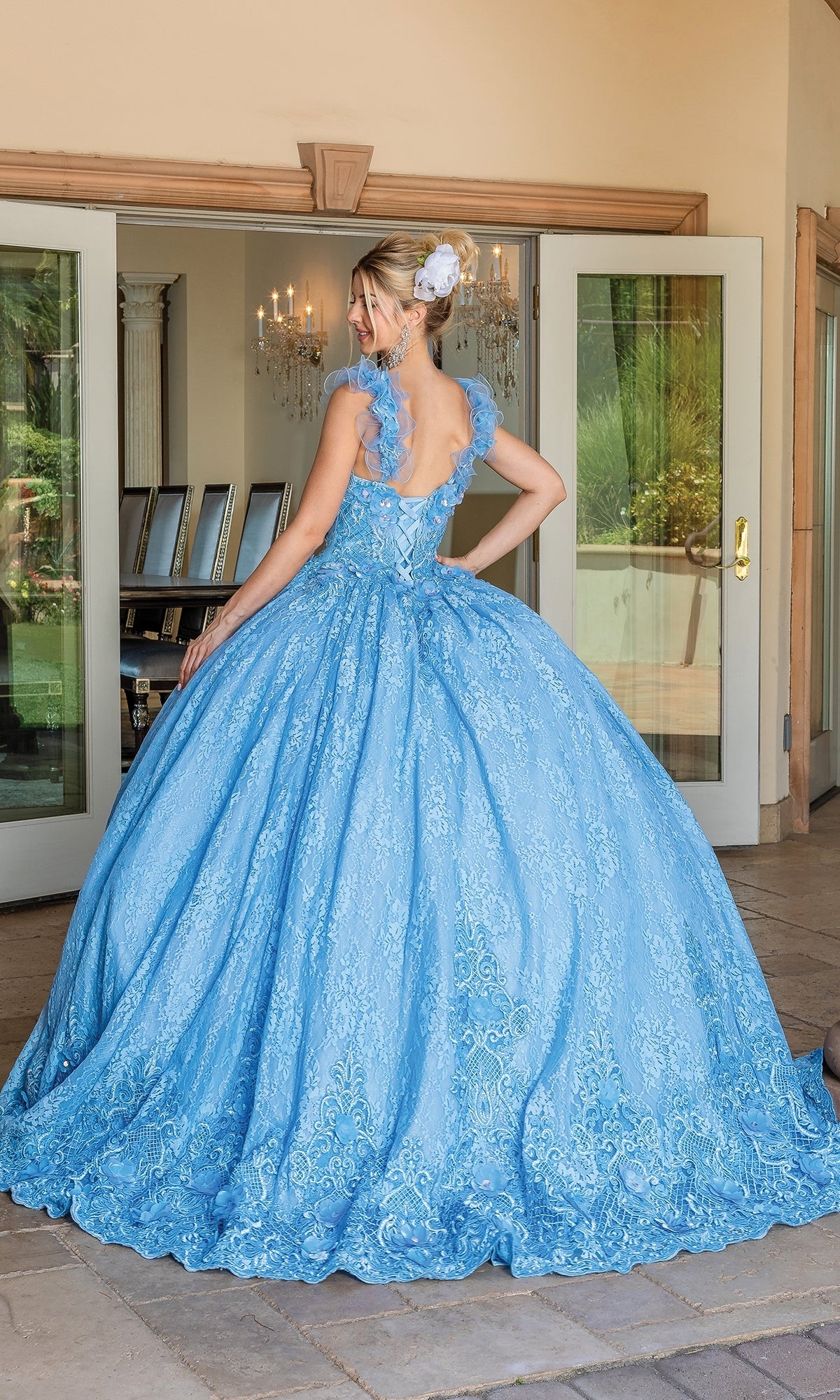 A woman with blonde hair in an updo wears the Quinceanera Dress 1721 By Dancing Queen, a light blue ball gown with a full skirt, lace details, and a sweetheart neckline, standing outdoors before open glass doors.